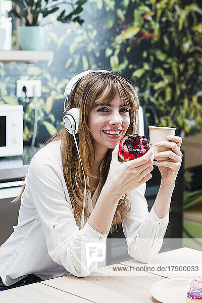 Lächelnde Geschäftsfrau mit Kopfhörern sitzt mit Einwegkaffeetasse und Donut in der Cafeteria