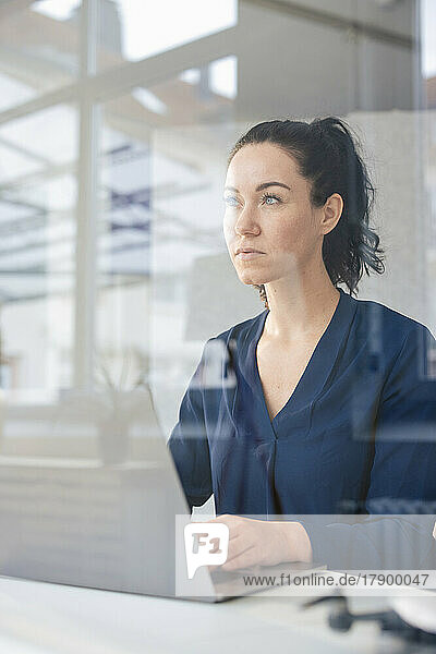Thoughtful businesswoman sitting with laptop seen through glass