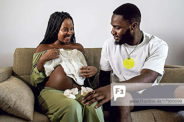 Pregnant woman showing baby clothing to man sitting on sofa at home