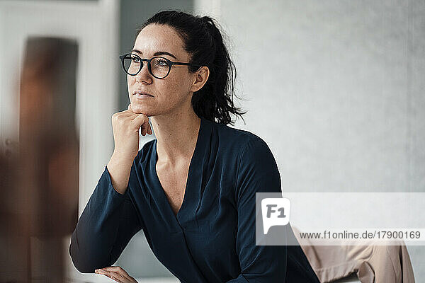 Nachdenkliche Geschäftsfrau sitzt mit der Hand am Kinn vor einer grauen Wand