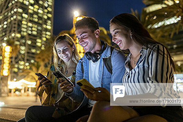 Happy young man sharing smart phone with friends at night