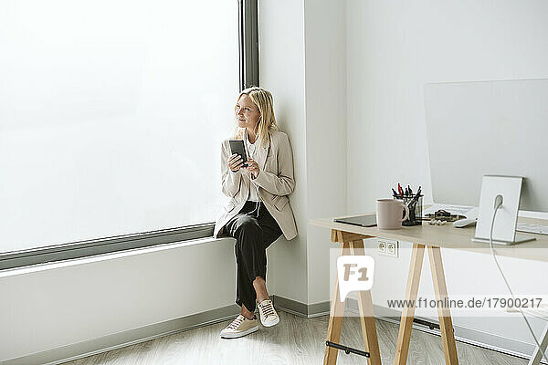 Geschäftsfrau sitzt auf der Fensterbank im Büro und hält ihr Mobiltelefon in der Hand