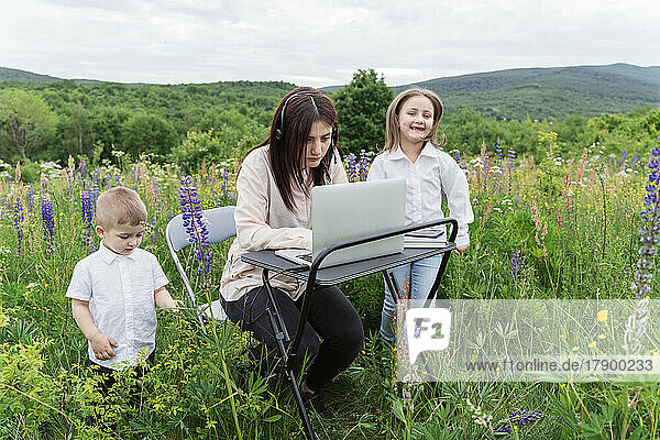 Mother working on laptop by playful children in meadow