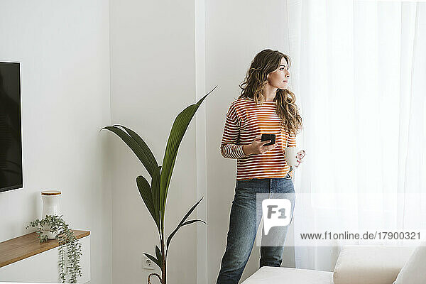 Thoughtful young woman holding coffee cup and mobile phone at home