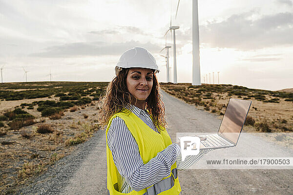 Smiling engineer with laptop standing at wind farm