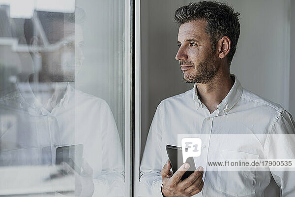 Thoughtful businessman holding mobile phone looking through window