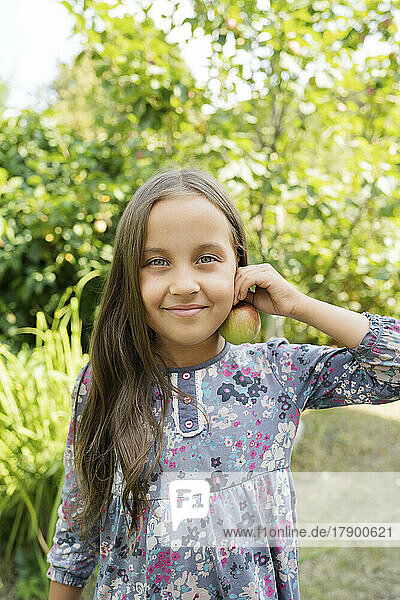 Smiling girl holding apple near ear in garden