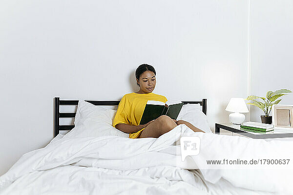 Frau im gelben T-Shirt sitzt auf dem Bett und liest ein Buch