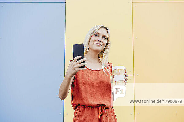 Smiling young woman holding mobile phone and disposable coffee cup in front of colorful wall