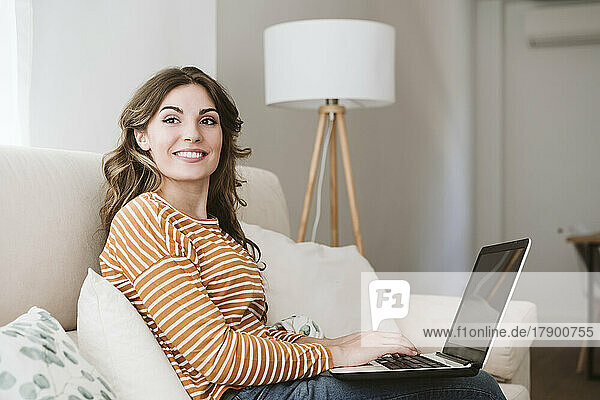 Lächelnde junge Frau  die zu Hause auf dem Sofa einen Laptop benutzt