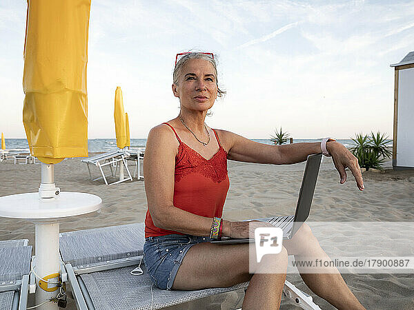 Nachdenkliche ältere Geschäftsfrau sitzt mit Laptop am Strand