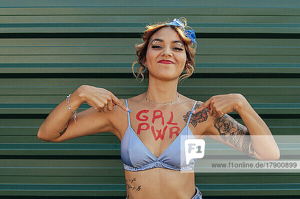 Selbstbewusste Frau zeigt Girl-Power-Text auf der Brust vor grüner Metallwand