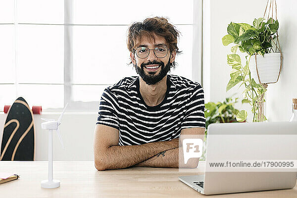 Lächelnder Mann sitzt neben Windkraftanlage und Laptop im Heimbüro
