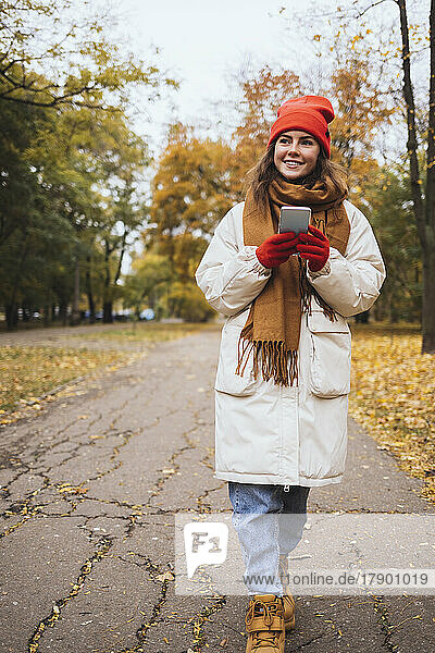 Lächelnde junge Frau mit Mobiltelefon geht auf Fußweg im Park