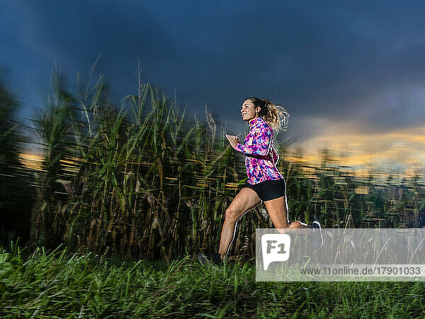 Smiling young sportswoman running on grass at dusk