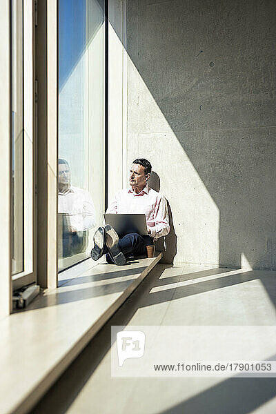 Businessman with laptop looking through window at office