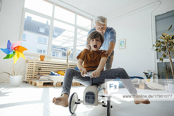 Happy grandfather pushing grandson sitting on toy car at home