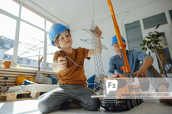 Boy imitating as engineer examining electricity pylon model with grandfather in background at home