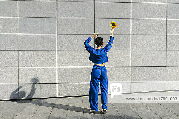Frau mit erhobenen Armen hält an einem sonnigen Tag eine Sonnenblume vor der Wand