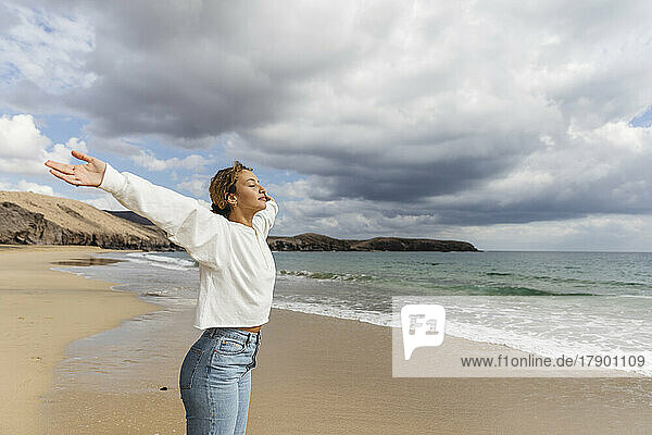 Junge Frau mit ausgestreckten Armen und geschlossenen Augen genießt den sonnigen Tag am Strand
