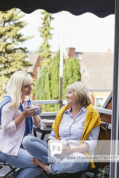 Smiling mother and daughter with coffee cups sitting together in balcony