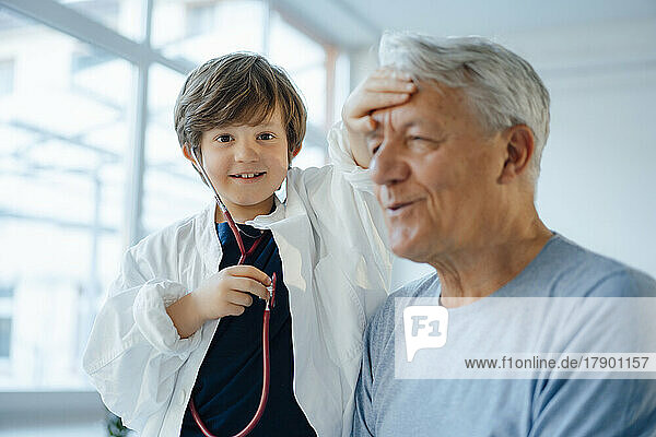 Lächelnder Junge imitiert als Arzt  der zu Hause die Temperatur seines Großvaters überprüft