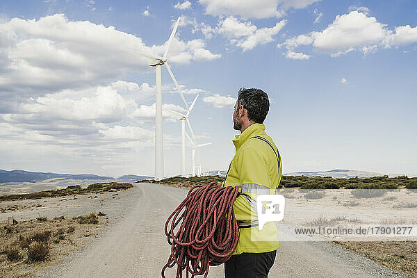 Technician holding rope looking at wind turbines standing on road at wind farm