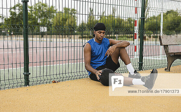 Young man with basketball leaning on fence sitting at sports court