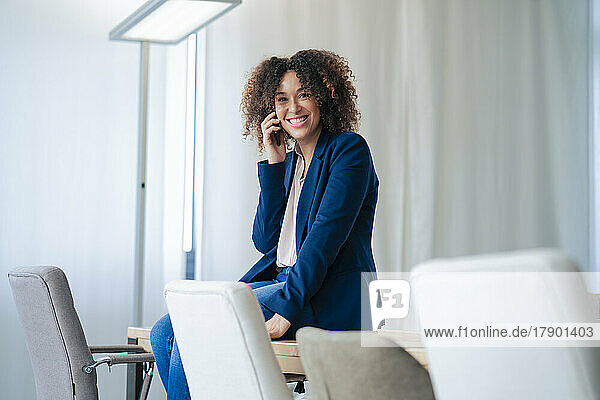 Happy businesswoman talking through mobile phone sitting on conference table in workplace