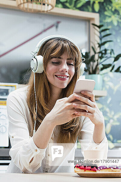 Smiling businesswoman with bangs text messaging through smart phone sitting in cafeteria