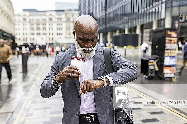 Senior businessman with disposable coffee cup checking time on wristwatch in city