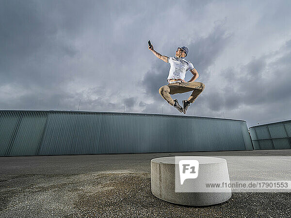 Young man jumping over concrete bench and taking selfie through mobile phone under cloudy sky