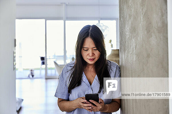 Mature woman using smart phone leaning on wall at home