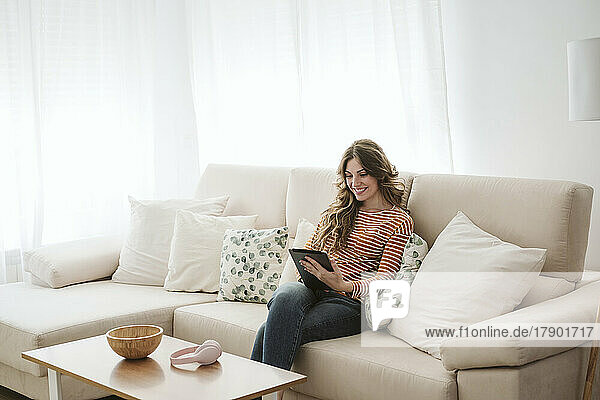 Lächelnde junge Frau nutzt digitales Tablet auf dem heimischen Sofa