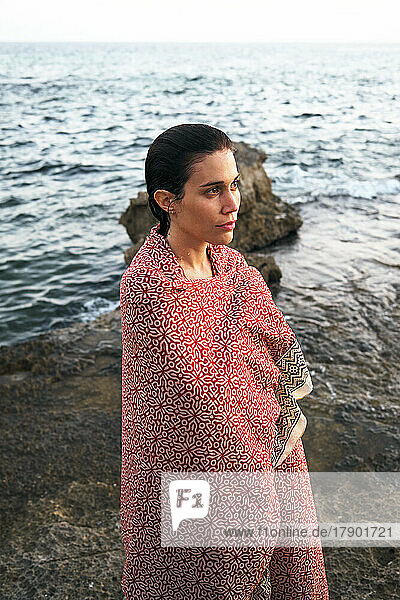 Contemplative woman wrapped in blanket standing on shore