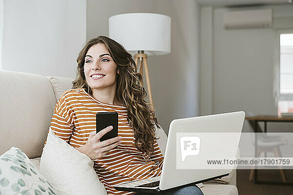 Smiling young woman with laptop and mobile phone on sofa at home