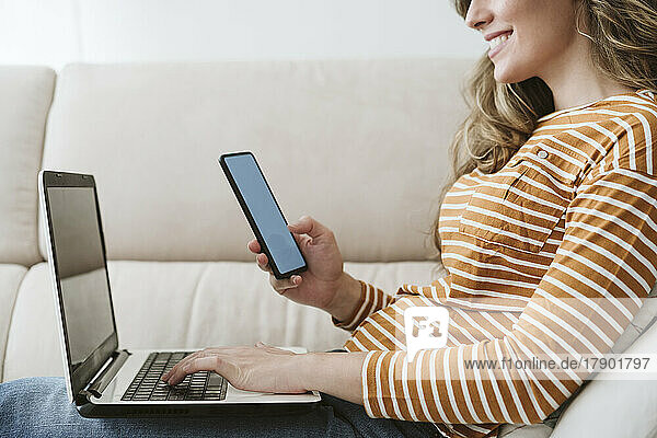 Woman using laptop and mobile phone on sofa