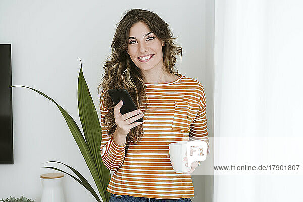 Portrait of smiling young woman holding coffee cup and mobile phone