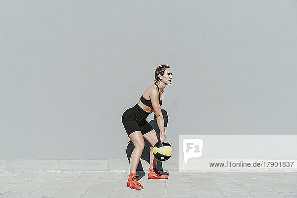 Woman with medicine ball exercising in front of gray wall on sunny day