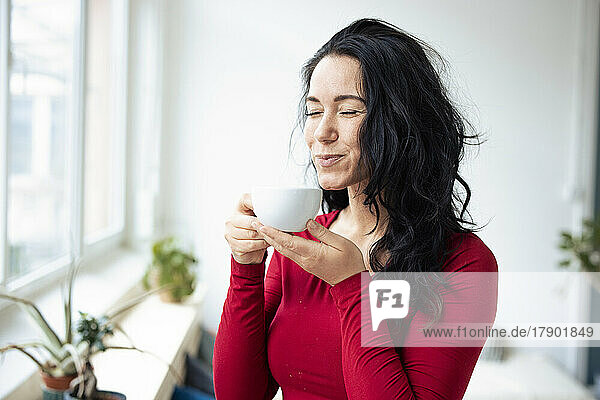 Woman with eyes closed holding coffee cup standing by window