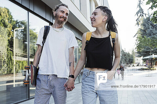 Happy young hipster couple holding hands walking at footpath