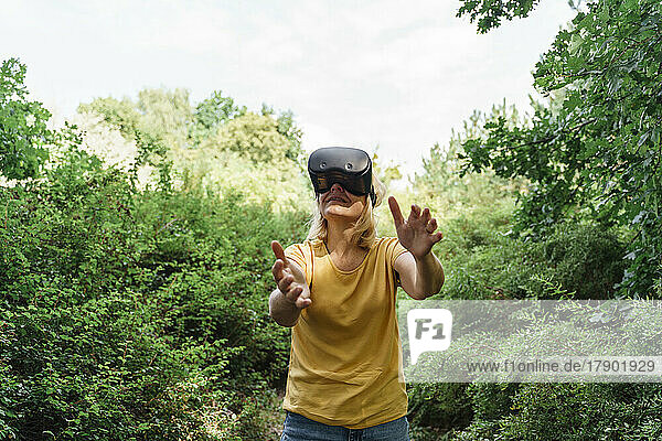 Mature woman gesturing with virtual reality simulators standing amidst plants