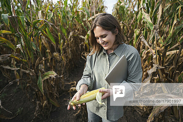 Smiling woman with laptop holding corncob in field