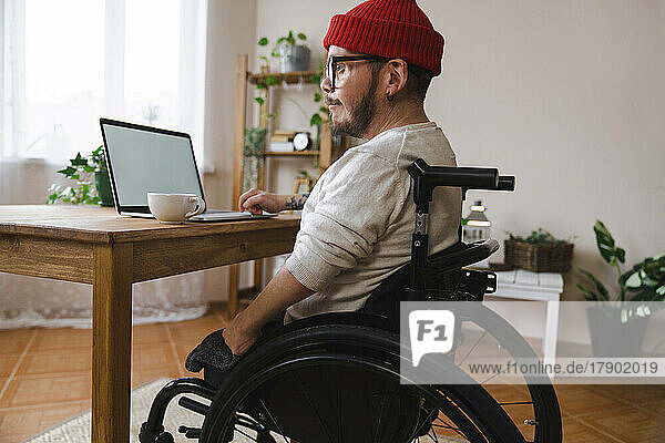 Nachdenklicher Mann im Rollstuhl mit Laptop zu Hause