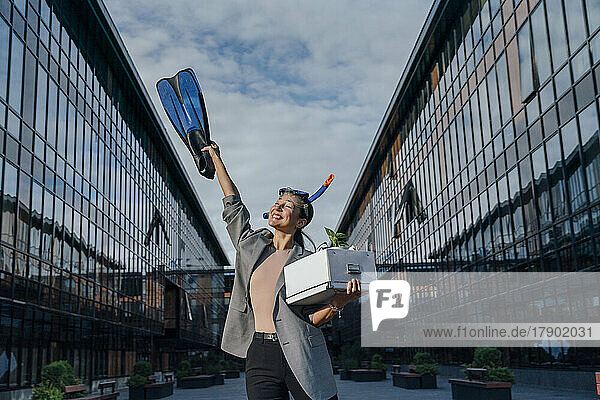 Happy businesswoman with diving flipper and box in front of office building