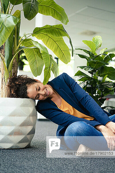 Smiling businesswoman resting head on potted plant in office