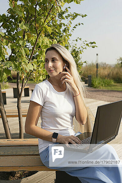 Smiling freelancer with laptop wearing in-ear headphones in park