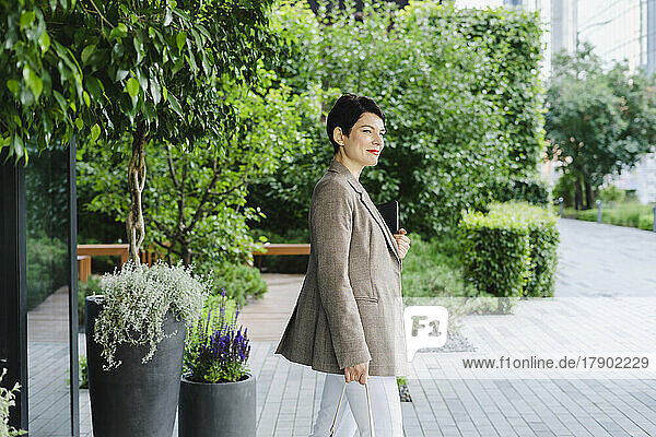 Smiling businesswoman with tablet PC standing by plants at office park