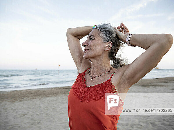Ältere Frau mit den Händen hinter dem Kopf steht am Strand