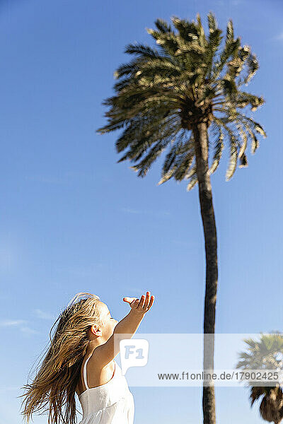 Girl with arm outstretched standing in front of palm tree on sunny day